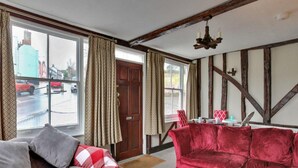 Una habitación con un sofá rojo, un sofá gris y una mesa de centro de madera. Hay una ventana con cortinas, una puerta y una lámpara de araña.