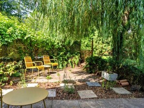 Eine Garten-Terrasse mit Tisch und Stühlen, umgeben von üppiger Bepflanzung und einer Trauerweide.