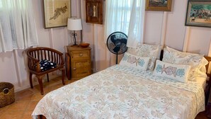 A bedroom with a bed, a chair, a nightstand, a fan, and a wall with framed pictures. - Beautiful Private Waterfront Estate, 2 Properties, 4 Bedrooms, West End Tortola (Tortola)
