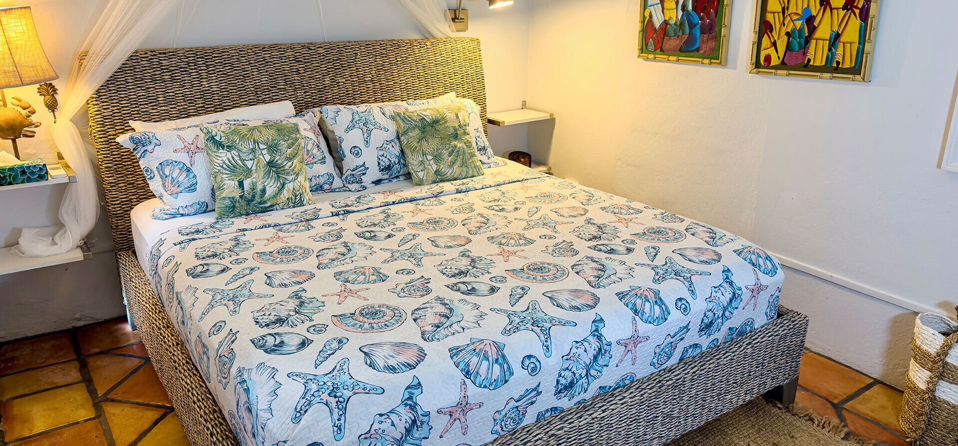 A bed with a tropical print bedspread, a wicker headboard, and a bedside table with a lamp.