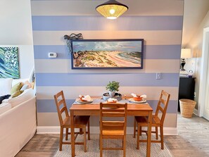 A dining area with a wooden table, four chairs, a painting of a beach scene, and a ceiling light fixture. - Serenity Now: Relax by the pool! Located in the Heart of the Village. (Ocracoke)