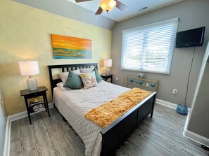 A bedroom with a bed, two bedside tables, a ceiling fan, a window with blinds, and a television on the wall. - Serenity Now: Relax by the pool! Located in the Heart of the Village. (Ocracoke)