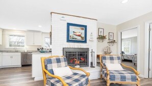 A room with a fireplace, two armchairs, a dining table, and a kitchen area. - Charming Seaside Retreat – Walk to the Beach in Rye, NH (Rye)