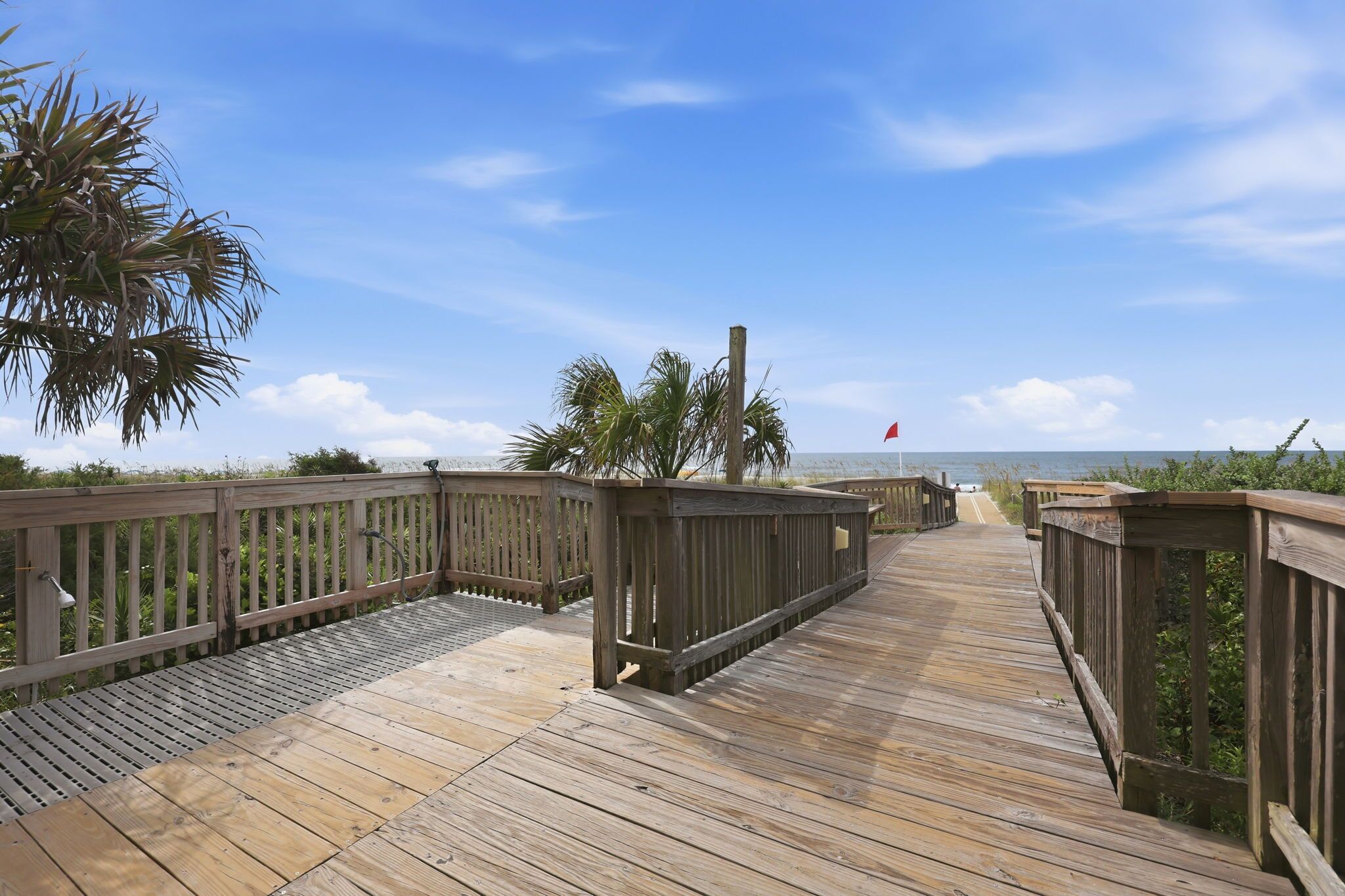 A wooden boardwalk leading to a beach with palm trees and a red flag.