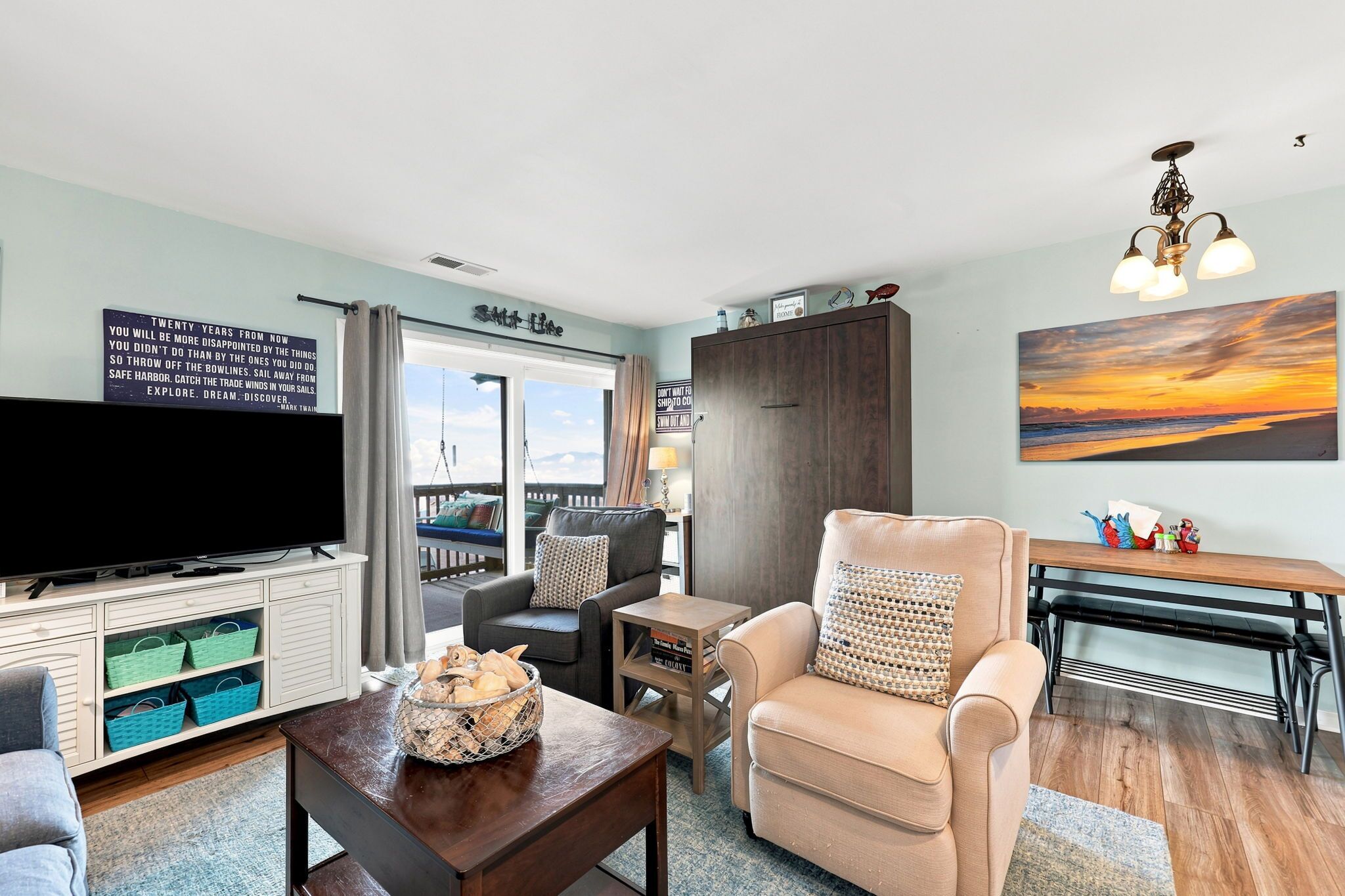 A living room with a television, a sofa, a chair, a coffee table, a bookshelf, and a view of the outdoors.