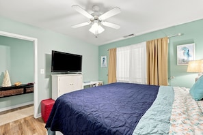 A bedroom with a bed, a television, a ceiling fan, and a window with curtains. - Atlantic Queen - Oceanfront Escape (Emerald Isle)