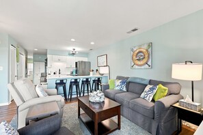 A modern living room with a grey sofa, a coffee table, and a kitchen in the background. - Atlantic Queen - Oceanfront Escape (Emerald Isle)