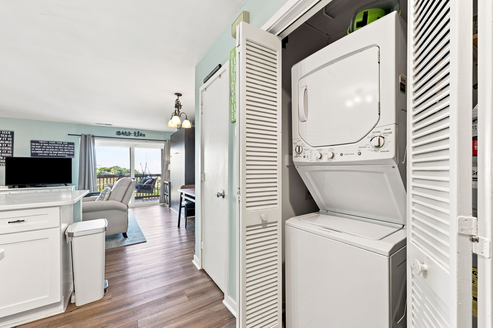 A laundry room with a front-loading washer and dryer, a small trash bin, and a door leading to a balcony with a view.
