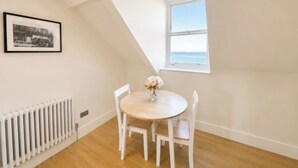 Een kamer met een ronde houten tafel, witte stoelen, een radiator, een lijst met een afbeelding en een raam met uitzicht op zee.