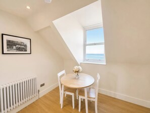 A room with a round wooden table, white chairs, a radiator, a framed picture, and a window with a view of the sea. - 123 On The Sea The Eaves (Weymouth)