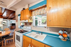 A compact kitchen with wooden cabinets, a built-in oven, a sink, and a window with a decorative curtain. - Seabright Surf Shack | Oak Island Beach Bungalow by Brunswick Vacation Rentals (Oak Island)