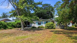 A single-story house with a front porch, surrounded by a grassy yard and trees. - Seabright Surf Shack | Oak Island Beach Bungalow by Brunswick Vacation Rentals (Oak Island)