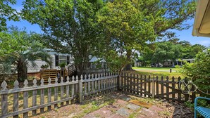 A wooden fence, a paved path, and a grassy area with trees. - Seabright Surf Shack | Oak Island Beach Bungalow by Brunswick Vacation Rentals (Oak Island)