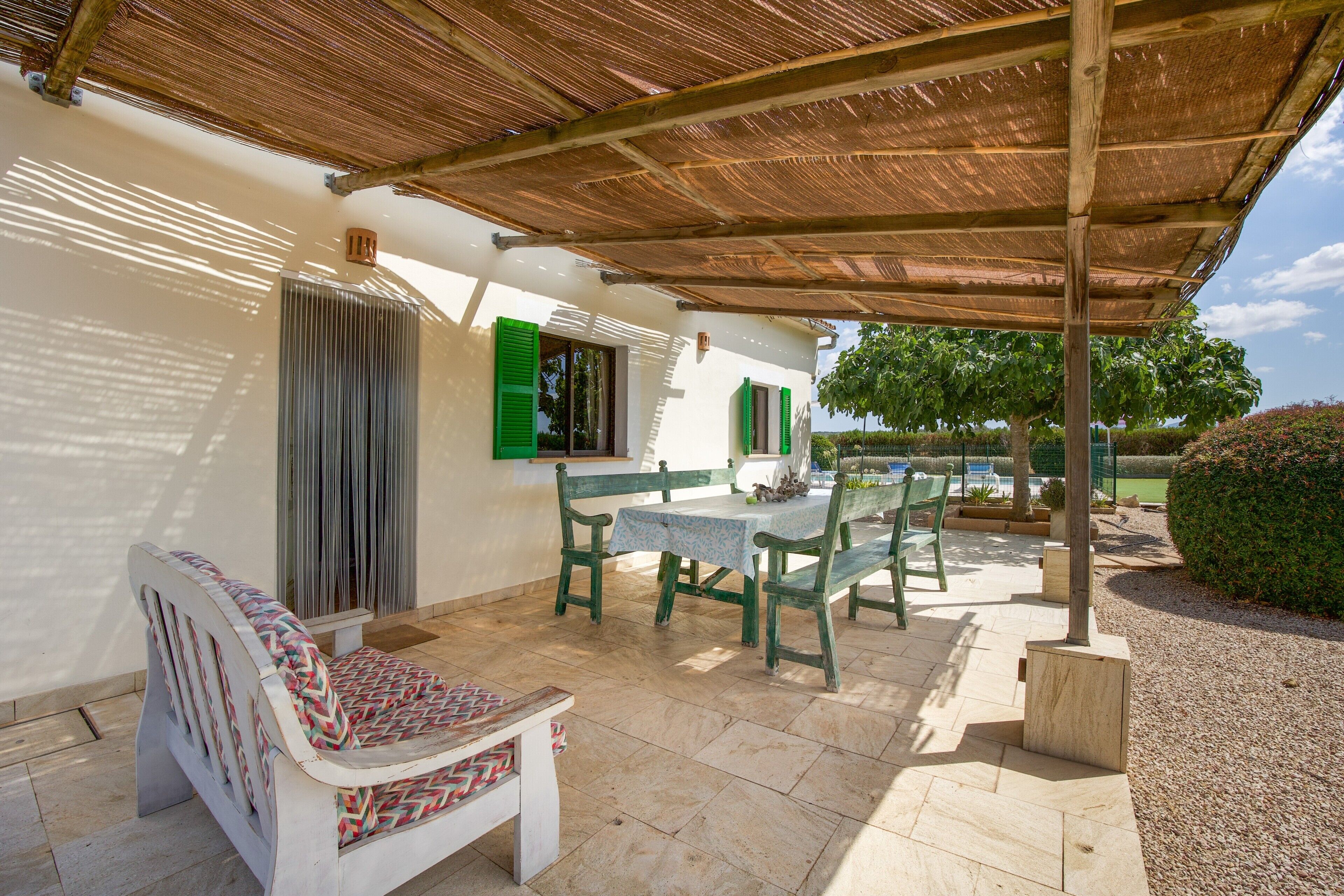 Een overdekt terras met een tafel, stoelen, een bankje en uitzicht op een zwembad en tuin.