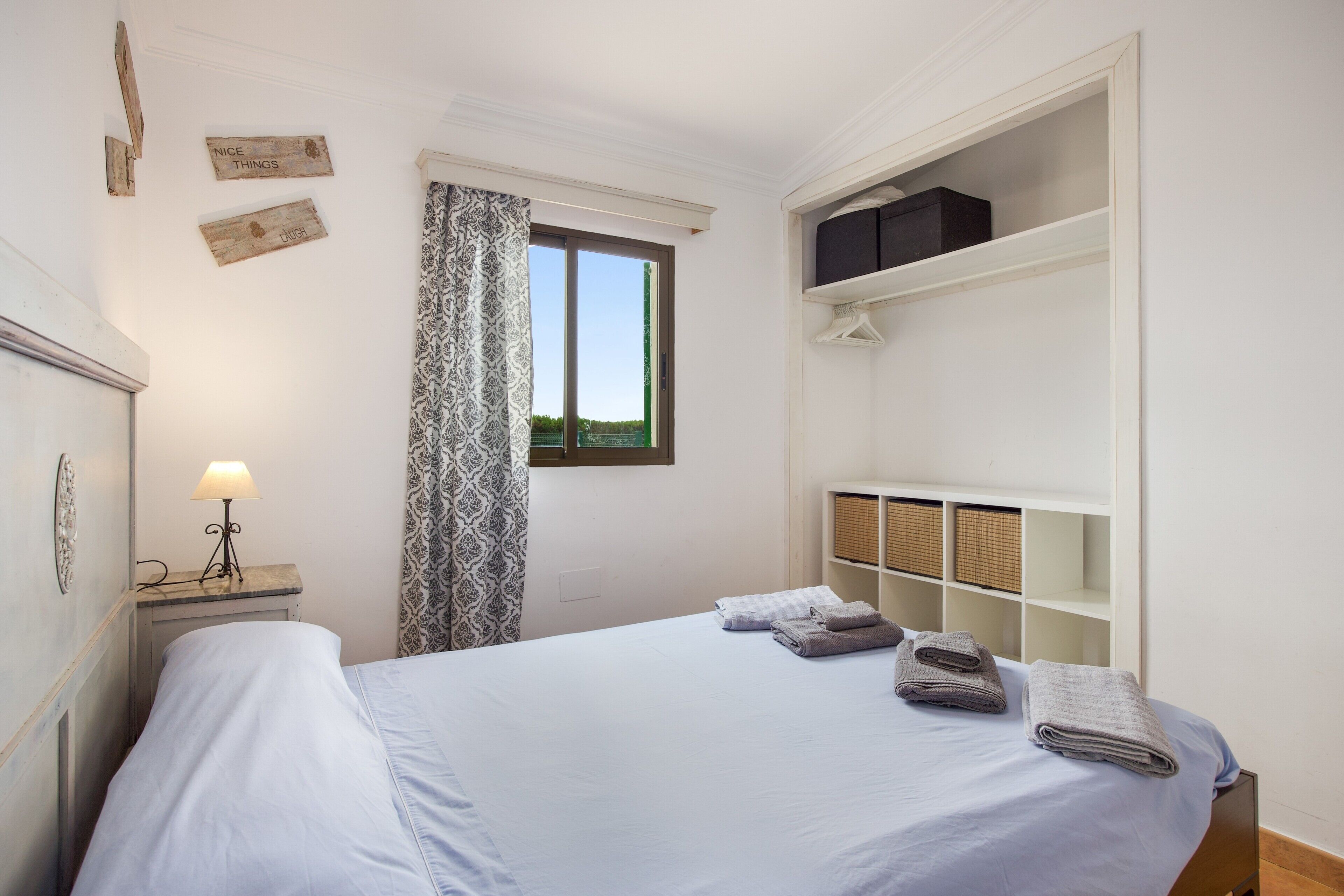 Un dormitorio con cama, mesita de noche con lámpara, ventana con cortinas y armario con estantes.