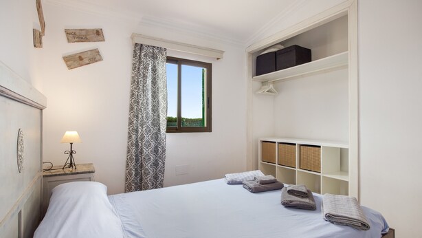 Een slaapkamer met een bed, een nachtkastje met een lamp, een raam met gordijnen en een kledingkast met planken.