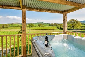 Ein Whirlpool mit Flasche und Glas, umgeben von einer Holzterrasse und einem Metallgeländer, mit Blick auf eine grüne Landschaft.