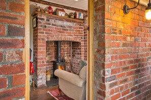 A cozy room with a brick fireplace, a comfortable armchair, and a shelf with various items. - Rustic Romantic Cottage in Llangadog (Llangadog)