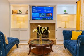 A living room with a television displaying 'Soecrum' on the screen, a fireplace, two blue sofas with yellow cushions, and a glass coffee table with a vase of flowers. - Executive 4BR Home Fully Furnished. CALL IT HOME 30+Day (Louisville)