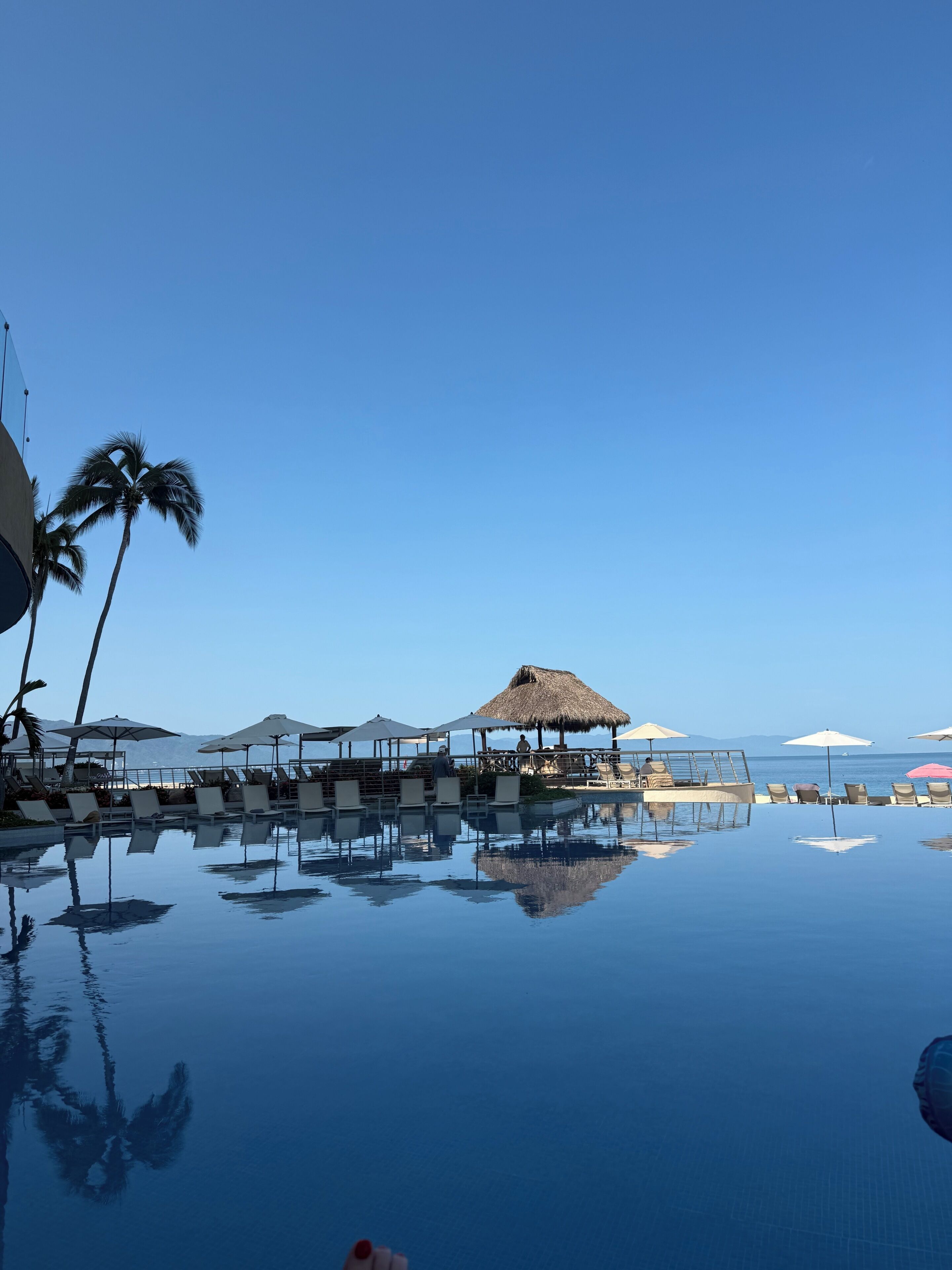 Cielo azul despejado, una piscina tranquila que refleja el entorno, y una estructura de techo de paja con asientos.