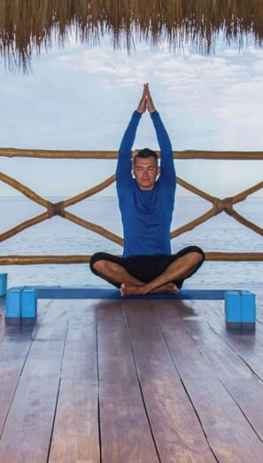 Una persona sentada en posición de meditación en una terraza de madera con barandilla y vista al agua.