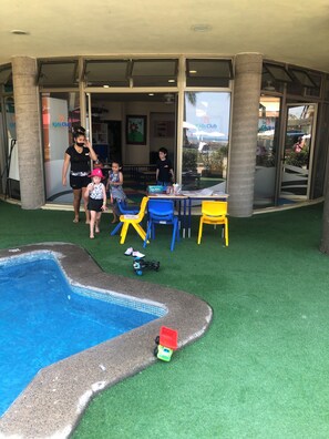 Ein Poolbereich mit Kunstgrras, ein Spielbereich für Kinder mit bunten Stühlen und ein sichtbares Schild für den Kids Club.