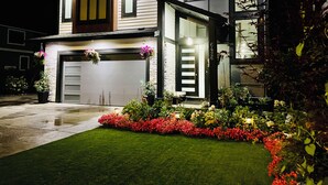 Een modern huis met twee verdiepingen, een garage, een verzorgd gazon en diverse bloeiende planten.