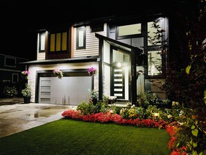 Uma casa moderna de dois andares, com garagem, gramado bem cuidado e diversas plantas floridas.