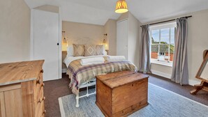Ein Schlafzimmer mit einem Bett, einer Holzkistenkommode, einem kleinen Holztisch, einem Fenster mit Vorhängen und einer Hängelampe.