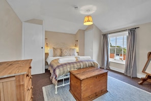 Ein Schlafzimmer mit einem Bett, einer Holzkistenkommode, einem kleinen Holztisch, einem Fenster mit Vorhängen und einer Hängelampe.