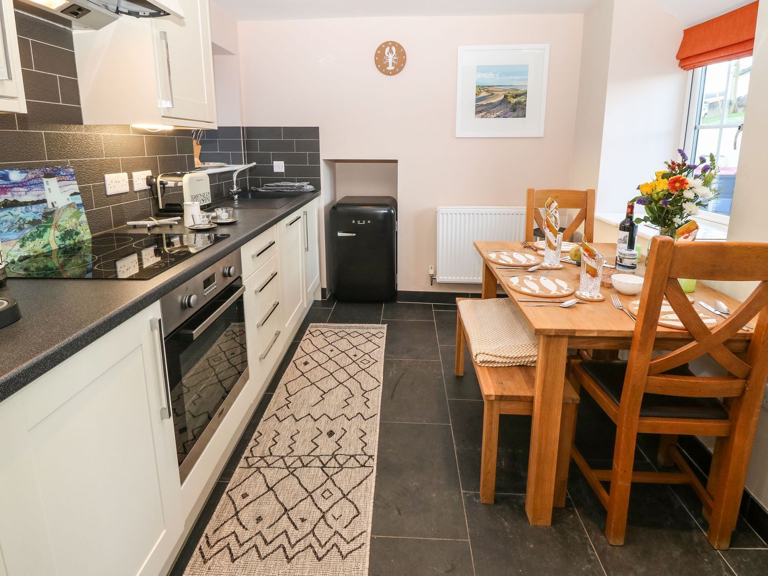 Una cocina funcional con un refrigerador negro, una mesa para dos personas y un tapete estampado en el suelo.