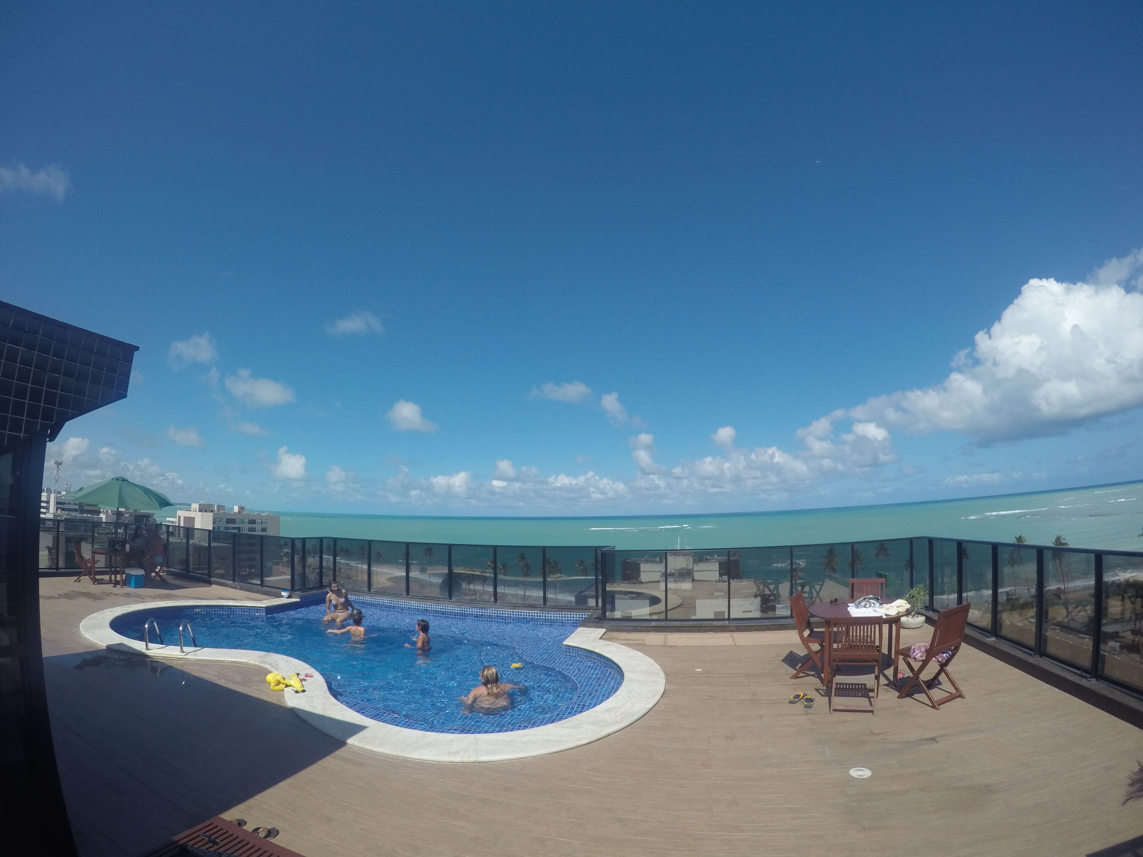 A rooftop pool area with a clear view of the ocean, surrounded by a glass barrier and furnished with outdoor seating.