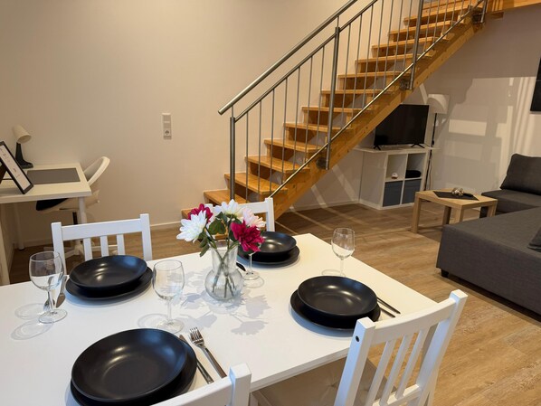 A dining area with a white table set for four, a vase with flowers, and a staircase in the background.