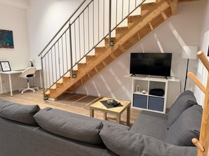 A modern living room with a wooden staircase, a grey sofa, a desk with a chair, and a television on a white stand.