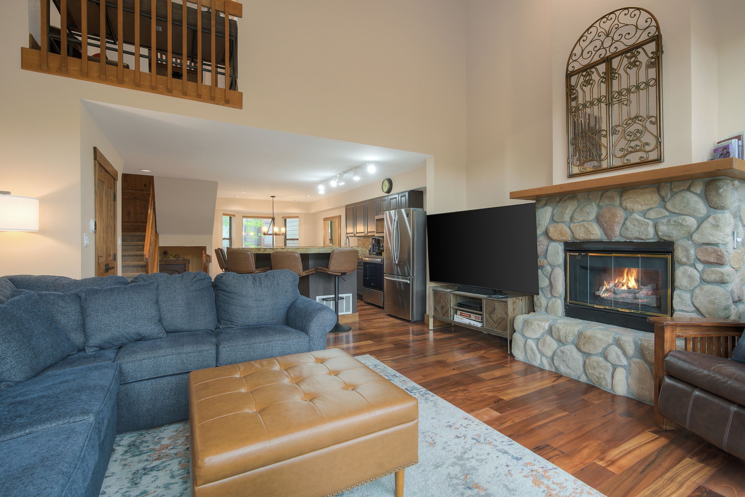 A modern living room with a sectional sofa, a leather ottoman, a stone fireplace, and a kitchen in the background.