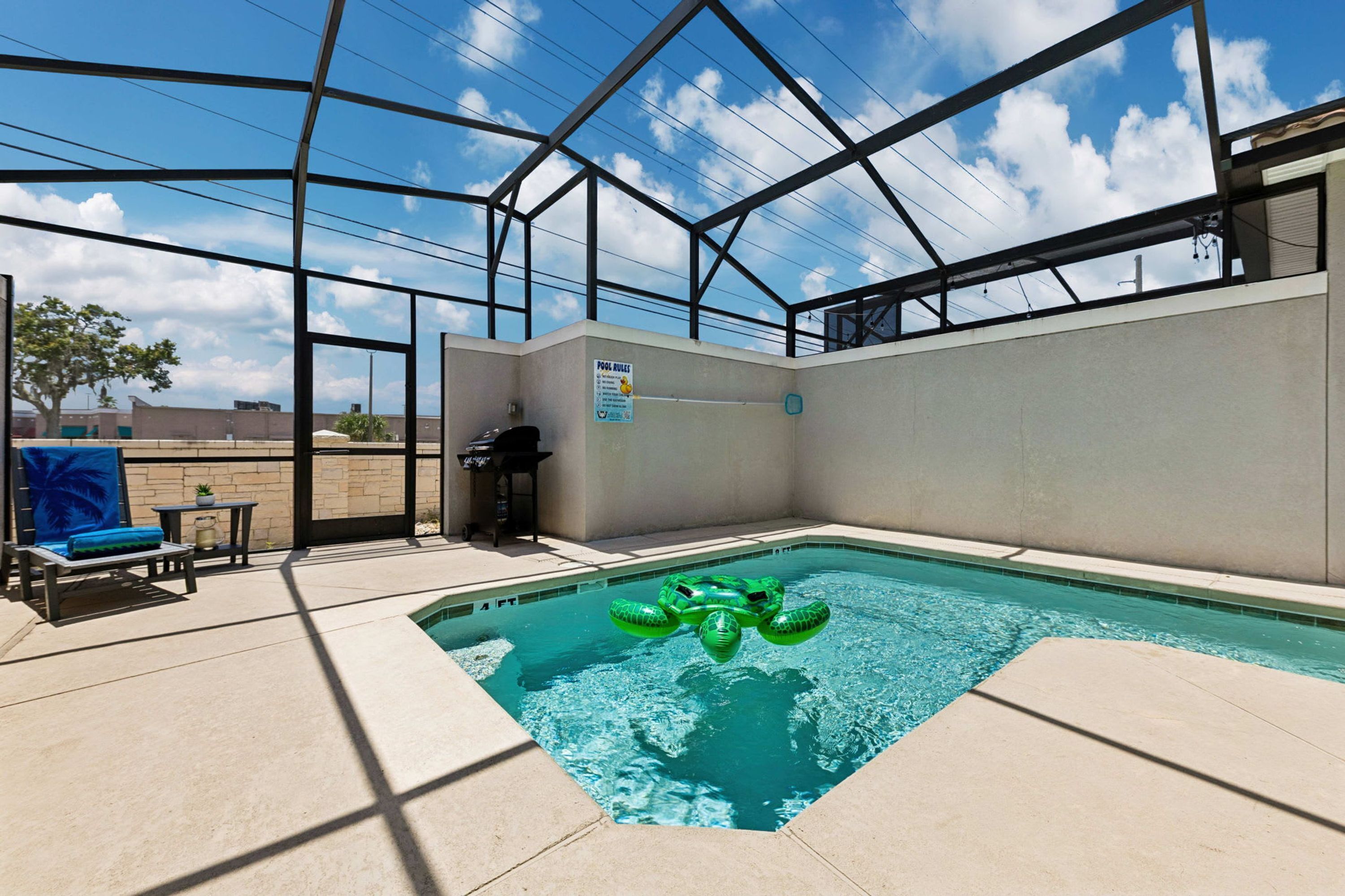 An indoor swimming pool with a green inflatable turtle float, a pool rule sign, and a patio area with lounge chairs and a table.