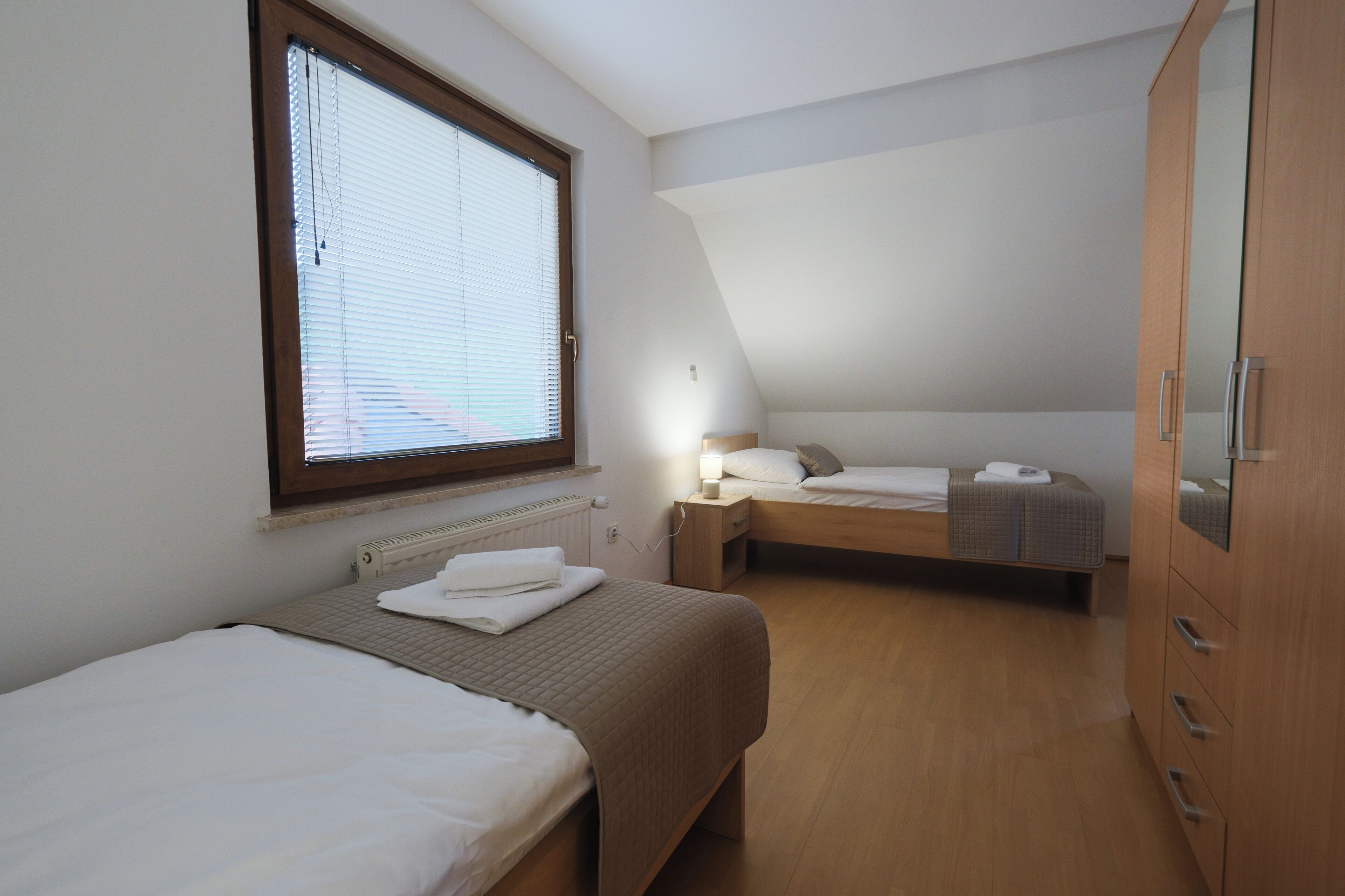 A bedroom with two beds, a wardrobe, a window with blinds, and a bedside table with a lamp.