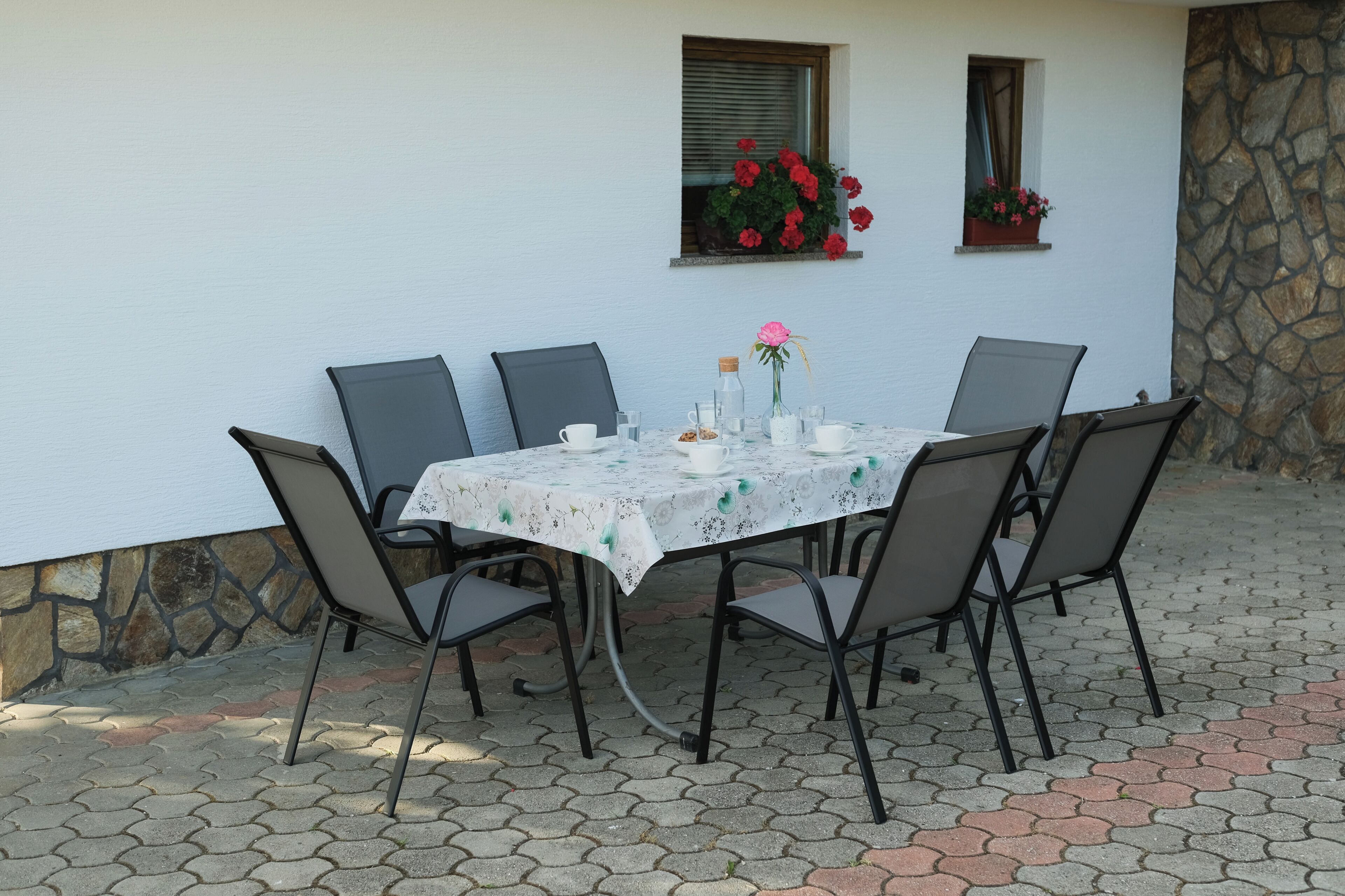 Een eethoek buiten met een tafel voor zes personen, omringd door stoelen, tegen een witte muur met een stenen fundering.