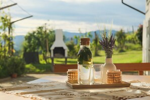 A table with a bottle, a glass, and a vase with lavender, set against a backdrop of a house and greenery.