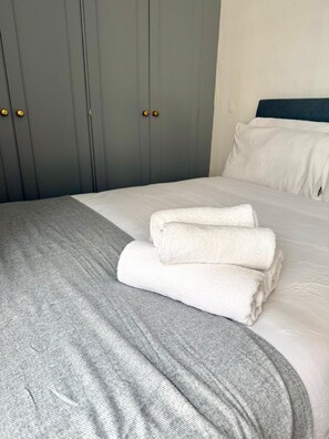 Ein ordentlich bezogenes Bett mit weißen Laken und einer grauen Decke, ein grauer Kleiderschrank mit goldfarbenen Griffen und ein weißes Kopfkissen auf dem Bett.