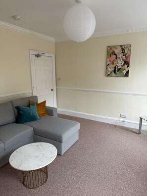 Ein Wohnzimmer mit einem grauen Ecksofa, einem weißen Couchtisch, einer weißen Deckenleuchte, einer weißen Tür und einem Blumenbild an der Wand.