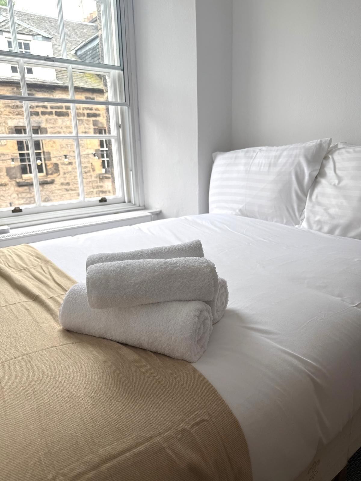 A neatly made bed with white linens and a beige blanket, a window with a view of a stone building, and a white wall.