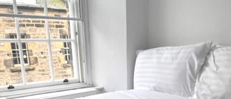 A neatly made bed with white linens and a beige blanket, positioned next to a window with a view of a stone building.