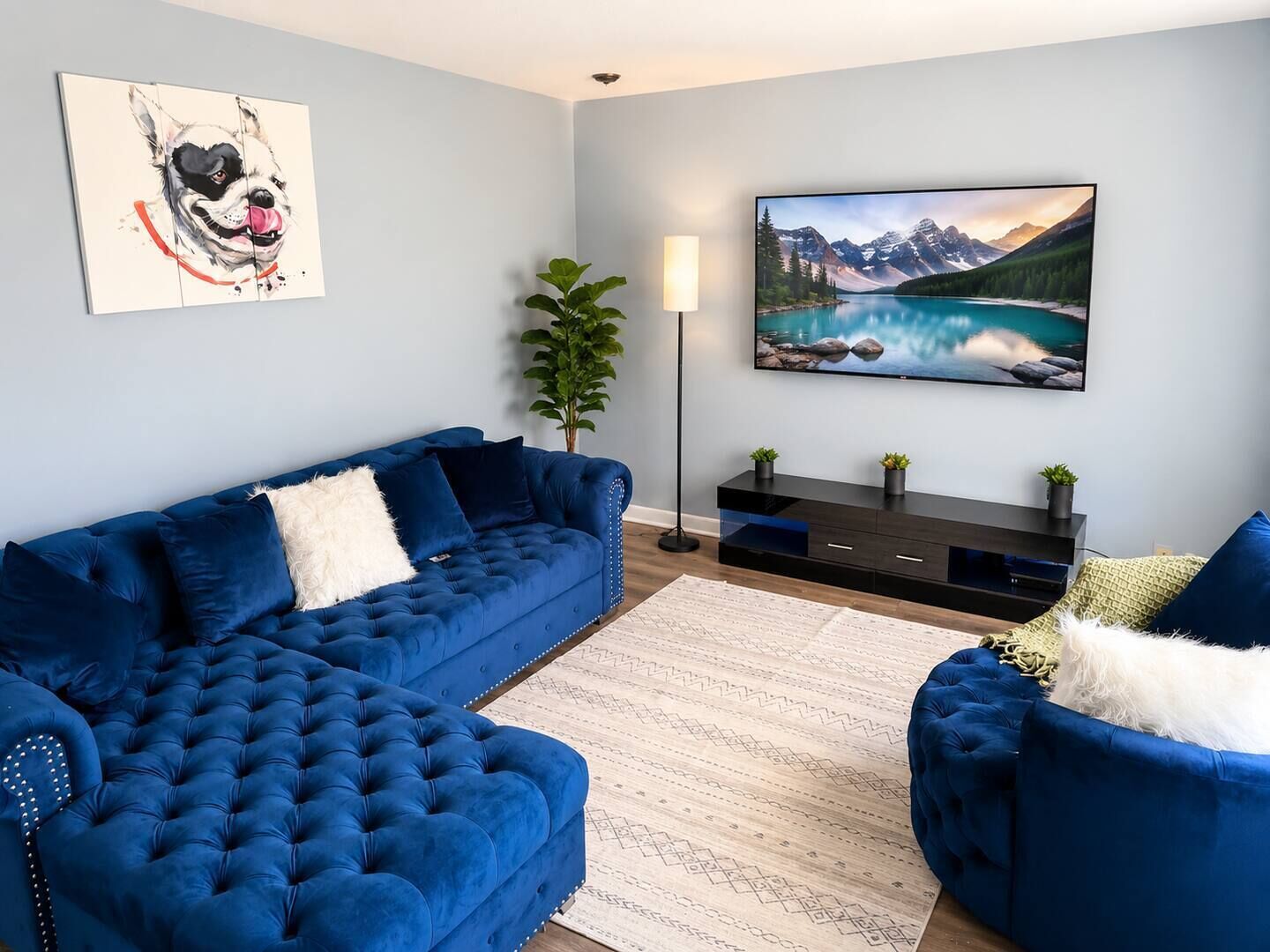 A living room with a blue sectional sofa, a flat-screen TV, a painting of a mountain landscape, and a potted plant.