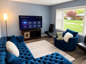 A living room with a flat-screen TV, two blue sofas, a patterned rug, and a window with a view of a green lawn and a tree. - Spacious 5BR Home for Families, Crews & Nurses 30+Day (Louisville)