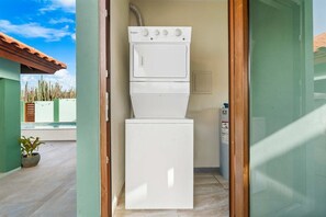A compact laundry room with a stacked washer and dryer, a potted plant, and a view of a pool and cacti. - 5BR Villa w/ Private Pool & Palapa, Noord Aruba (Noord)