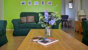 Ein Wohnzimmer mit einem grünen Sofa, einem Couchtisch mit Zeitschriften, einer Vase mit Blumen und einer grünen Wand mit gerahmten Bildern.