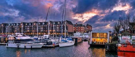 A marina with multiple boats docked, a row of buildings in the background, and a small structure with a sign.
