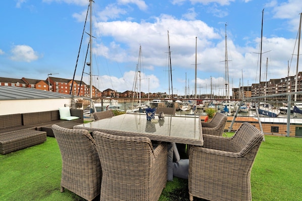 A rooftop terrace with a glass table and wicker chairs, overlooking a marina with sailboats and residential buildings.