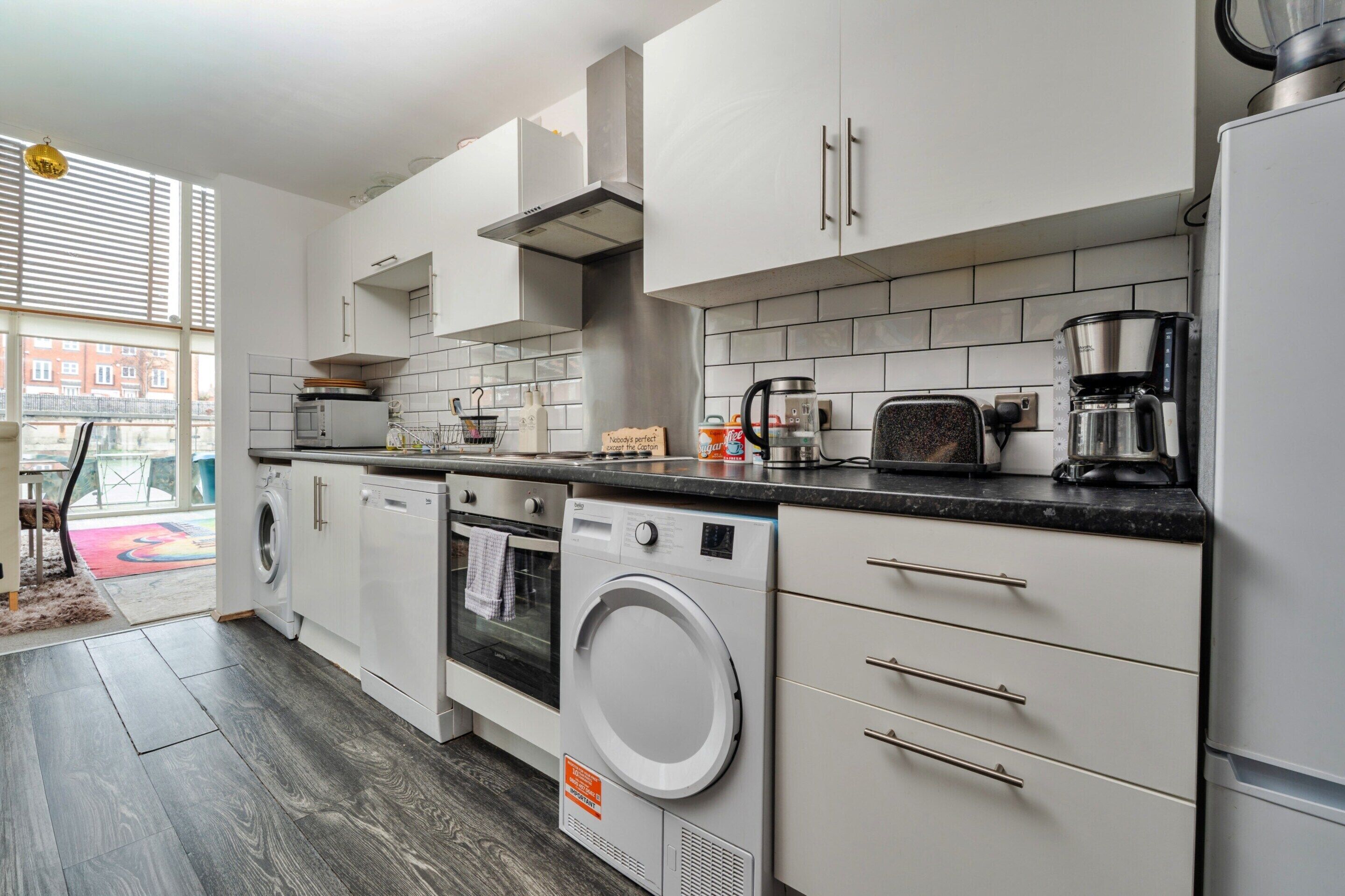 Una cocina moderna con armarios blancos, horno empotrado, lavadora y cafetera.